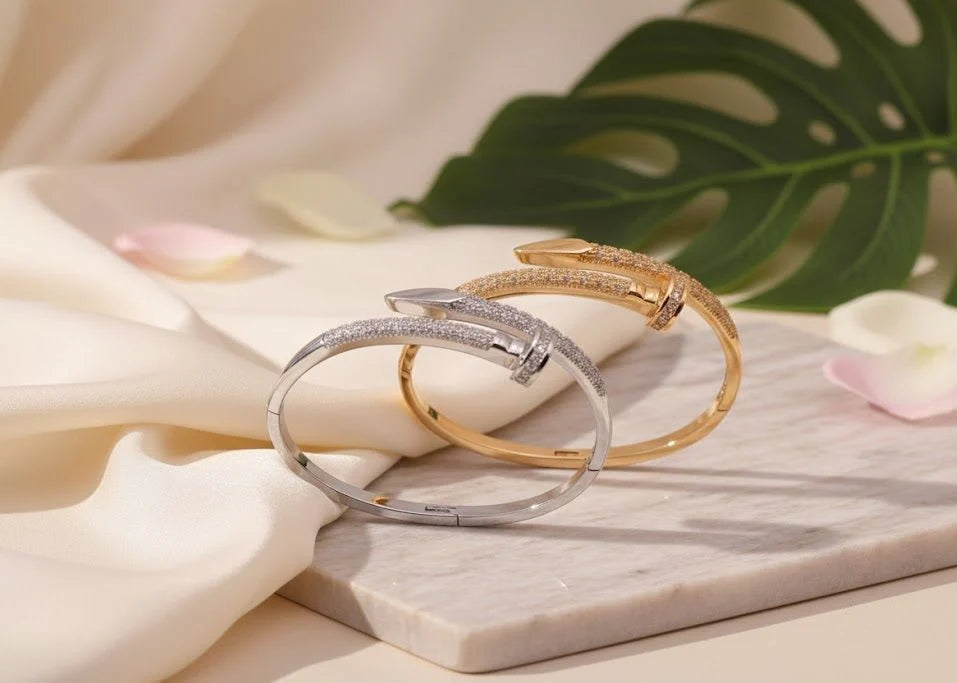 Two bracelets, one silver and one gold, on a marble surface with flowers and leaves in the background.