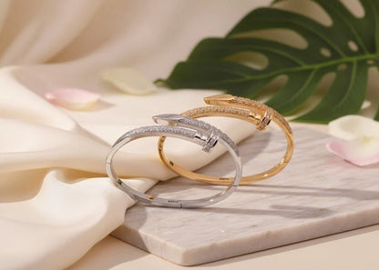 Two bracelets, one silver and one gold, on a marble surface with flowers and leaves in the background.